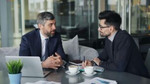 Dois homens sentados frente a frente em uma mesa, conversando em um ambiente interno profissional. Um deles gesticula enquanto fala, e o outro escuta atentamente, sugerindo uma reunião de negócios ou negociação. O cenário é neutro, com iluminação suave e foco na interação entre os dois.