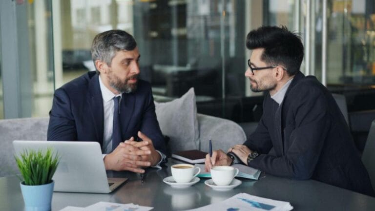 Dois homens sentados frente a frente em uma mesa, conversando em um ambiente interno profissional. Um deles gesticula enquanto fala, e o outro escuta atentamente, sugerindo uma reunião de negócios ou negociação. O cenário é neutro, com iluminação suave e foco na interação entre os dois.