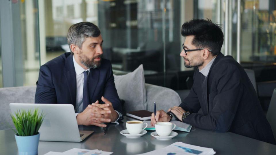 Dois homens sentados frente a frente em uma mesa, conversando em um ambiente interno profissional. Um deles gesticula enquanto fala, e o outro escuta atentamente, sugerindo uma reunião de negócios ou negociação. O cenário é neutro, com iluminação suave e foco na interação entre os dois.