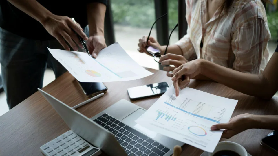 Esta imagem captura uma dinâmica de reunião de equipe ou auditoria financeira sob um ângulo superior focado na mesa de trabalho
