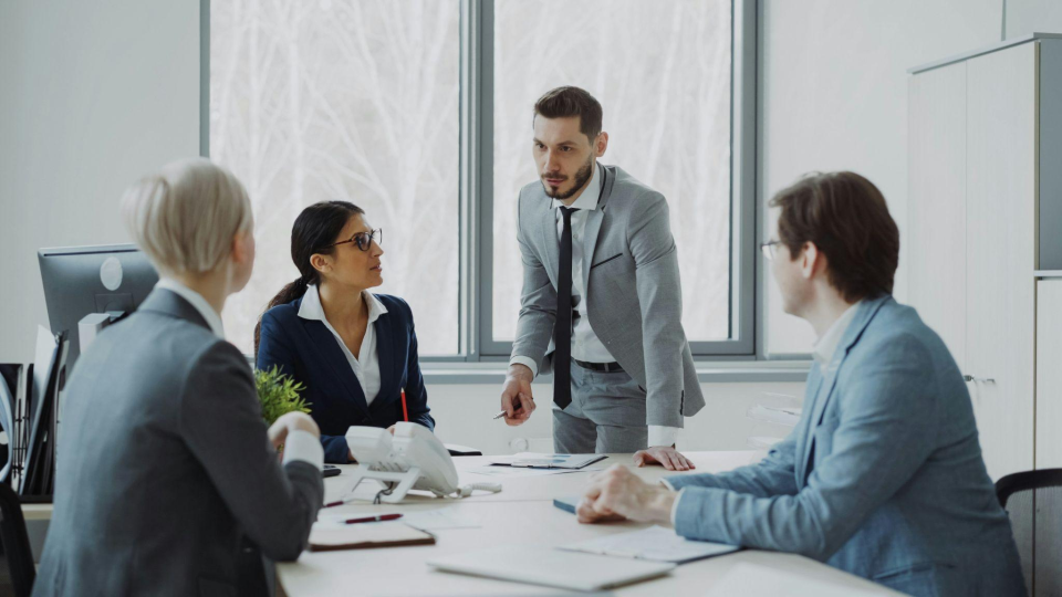 A imagem retrata uma reunião corporativa tensa ou de alinhamento estratégico conduzida por um líder em uma sala de escritório.