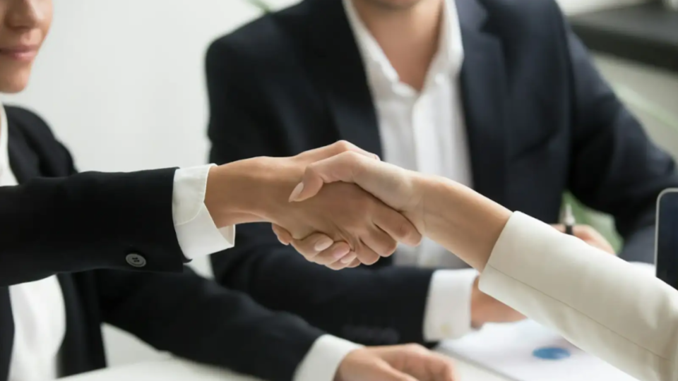 Dois profissionais em uma reunião corporativa fazem aperto de mãos sobre uma mesa, com escritório ao fundo, sugerindo acordo comercial.