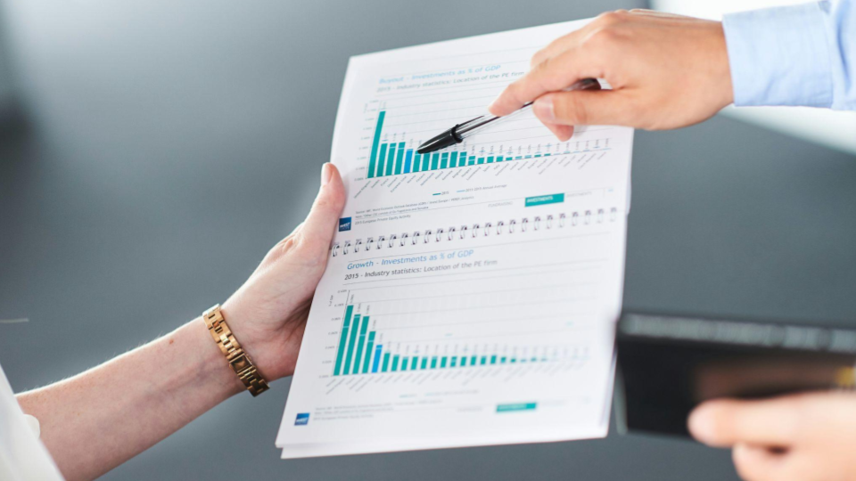 Mãos analisando um relatório financeiro com gráficos de barras e dados, em ambiente corporativo, durante uma reunião de negócios.