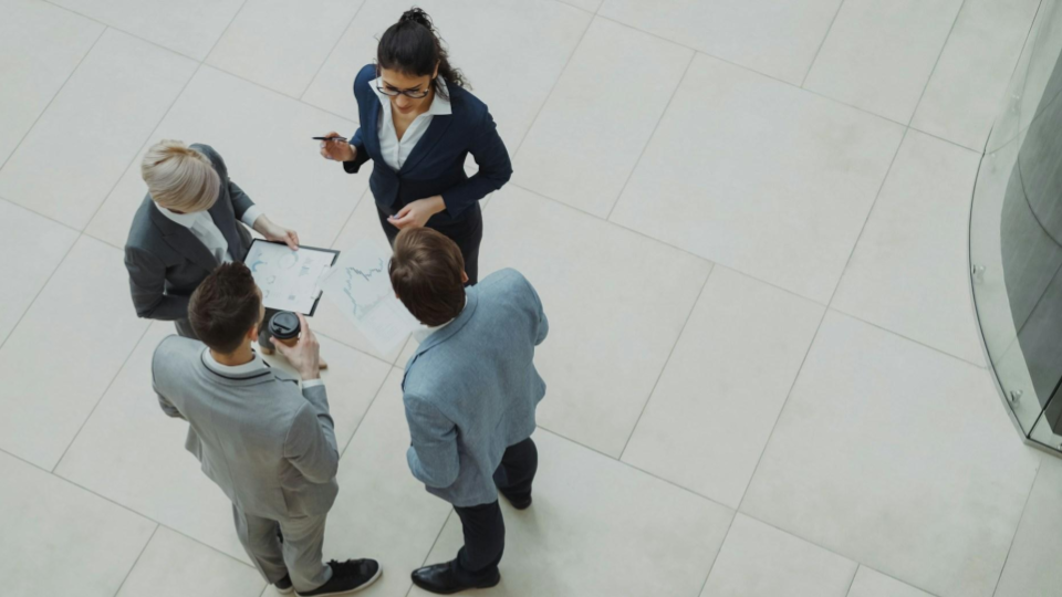 Equipe reunida em ambiente corporativo, com uma mulher analisando informações em papel e outras pessoas observando, em uma conversa de trabalho no escritório