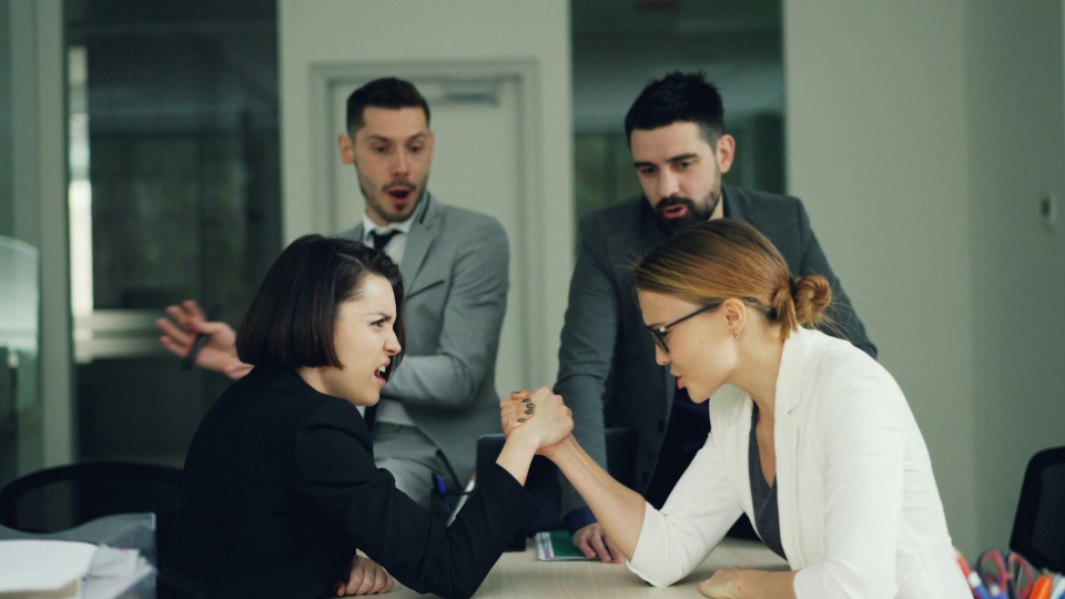 Equipe em um escritório, com dois homens no fundo e duas mulheres na frente, disputando uma queda de braço, imagem que ilustra um contexto de concorrência empresarial.