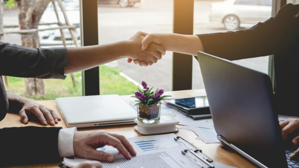 A foto mostra duas pessoas apertando as mãos em uma mesa de reunião, com notebook, documentos e um pequeno arranjo de flores ao centro, em ambiente corporativo com luz natural.