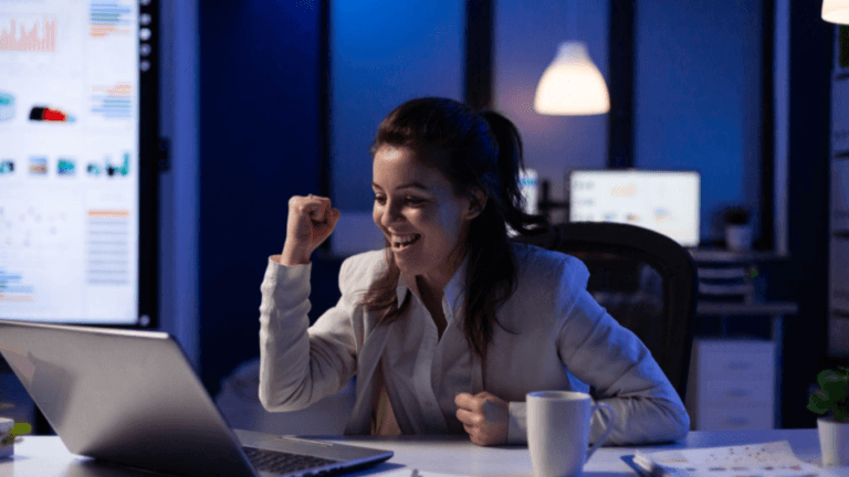 A imagem mostra uma jovem mulher profissional a comemorar um sucesso ou uma boa notícia à frente do seu computador portátil, num escritório moderno durante a noite.