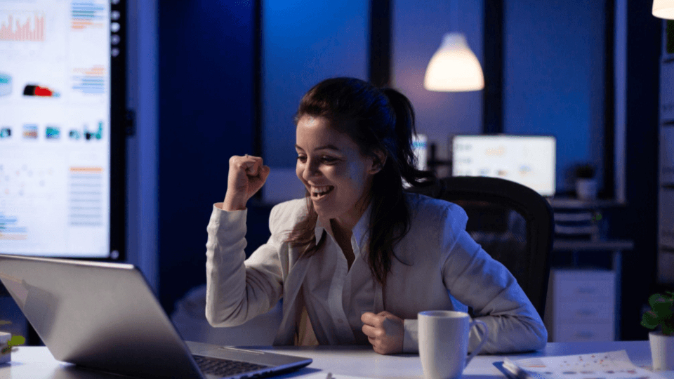 A imagem mostra uma jovem mulher profissional a comemorar um sucesso ou uma boa notícia à frente do seu computador portátil, num escritório moderno durante a noite.