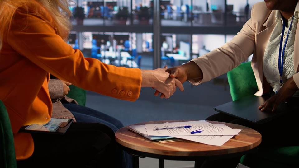A imagem retrata o aperto de mãos entre duas executivas em uma reunião de negócios, selando um acordo em um ambiente corporativo.
