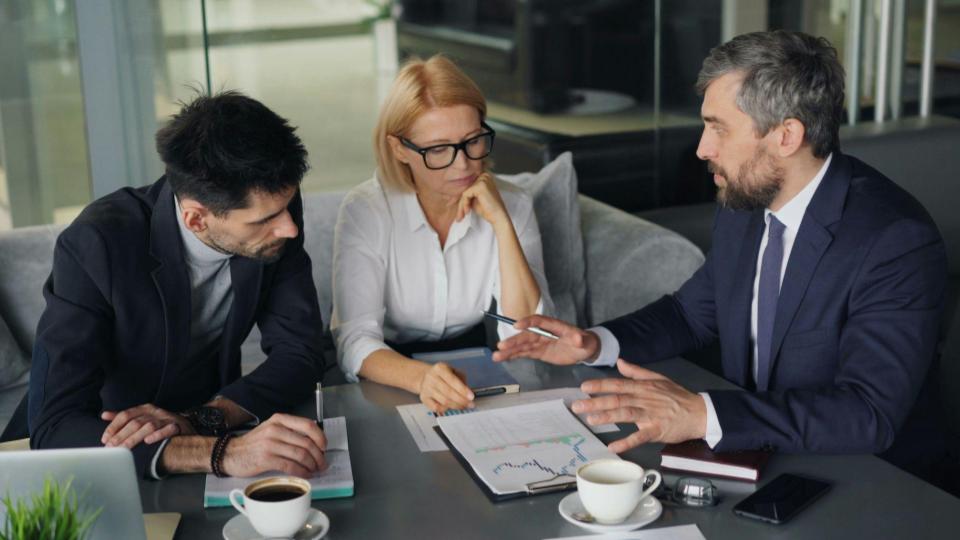 Três profissionais reunidos ao redor de uma mesa de centro com papéis, notebooks e xícaras de café, enquanto o homem à direita explica um ponto gesticulando com as mãos.