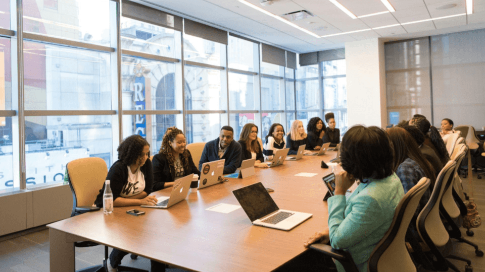 Uma equipa diversificada reunida em redor de uma grande mesa com portáteis numa sala corporativa iluminada.