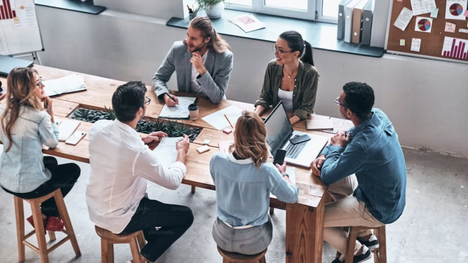 Vista superior de uma equipe corporativa de seis pessoas reunida ao redor de uma grande mesa de madeira rustica.