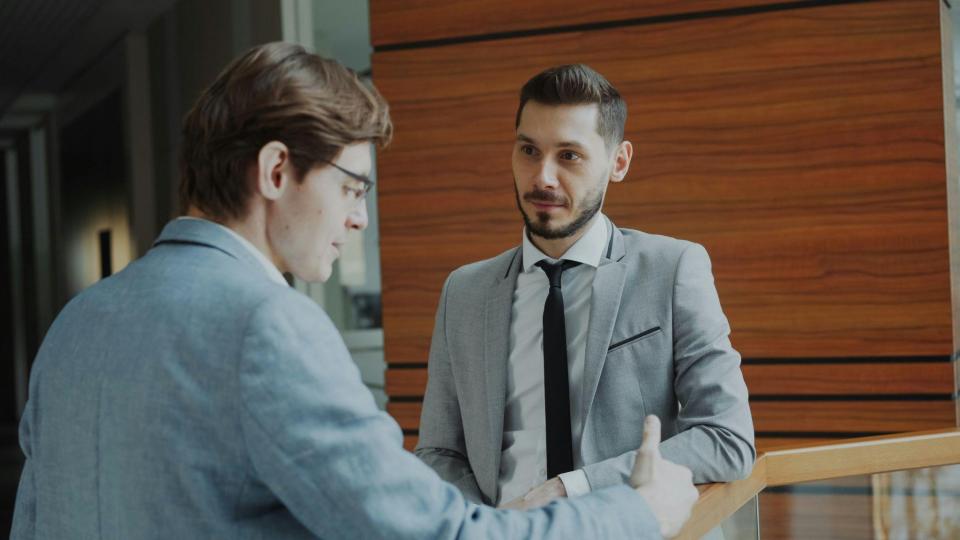 A imagem mostra dois homens em trajes de negócios conversando em um ambiente corporativo moderno.