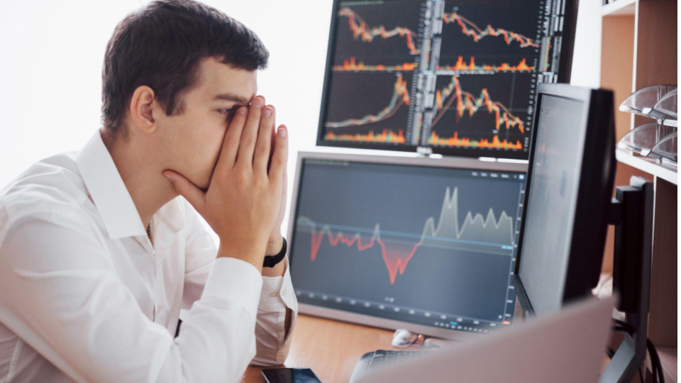 A imagem mostra um jovem profissional num ambiente de trabalho, visivelmente tenso ou sobrecarregado enquanto monitoriza o mercado financeiro.